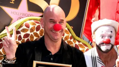 Guy Laliberté attends the Ceremony honouring him with a star on the Hollywood Walk of Fame on 22 November. He is wearing a red clown’s nose and has his hands in a V- sign.  To his left is a male clown with a white face paint, red nose, and a black beard and moustache painted into his face. The Hollywood Star is on a plaque behind them and Laliberté is holding a picture frame with the star inside.